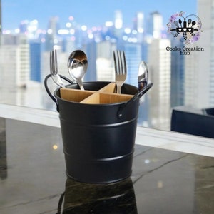 Puede incluir: Porta cubiertos de metal negro con un divisor de madera. El porta cubiertos tiene dos asas y está lleno de tenedores y cucharas.