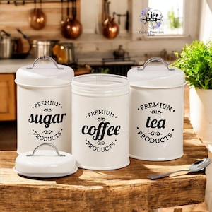 Może przedstawiać: Trzy białe metalowe pojemniki z czarnymi napisami i pokrywkami. Pojemniki są oznaczone napisami "PREMIUM. sugar PRODUCTS", "PREMIUM. coffee PRODUCTS" i "PREMIUM. tea PRODUCTS".
