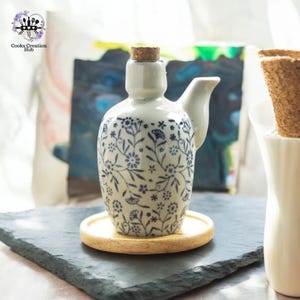 Może przedstawiać: Biała ceramiczna butelka z niebieskim wzorem kwiatowym i korkowym korkiem. Butelka ma dzióbek i uchwyt. Stoi na drewnianym podkładce na czarnej powierzchni z łupka.