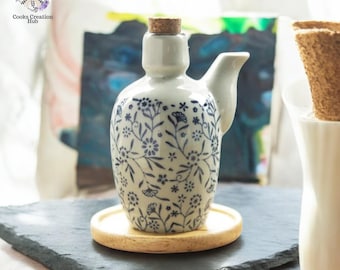 May include: A white ceramic bottle with a blue floral pattern and a cork stopper. The bottle has a spout and a handle. It is sitting on a wooden coaster on a black slate surface.