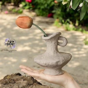 May include: A small, grey, textured ceramic vase with a single orange tulip inside. The vase has a handle and a rounded base.