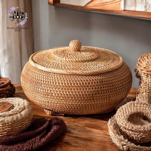 Könnte beinhalten: Ein großer, runder, geflochtener Korb mit Deckel. Der Korb ist aus natürlichen Materialien gefertigt und hat eine hellbraune Farbe. Der Korb steht auf einer Holzoberfläche.