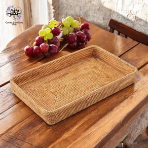 Puede incluir: Una bandeja rectangular de ratán tejido sobre una mesa de madera, con un racimo de uvas rojas y verdes en el fondo. La bandeja es de color marrón claro y tiene una superficie texturizada. El texto "Cooks Creation Hub" está en la esquina superior izquierda.