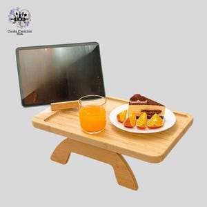 Puede incluir: Una bandeja de madera con una tableta, un vaso de zumo de naranja, una porción de pastel y rodajas de naranja en un plato blanco. La bandeja está sostenida por dos patas de madera.