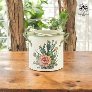 Puede incluir: Porta cubiertos de metal blanco con ribete verde y un diseño de cactus y flores. El porta cubiertos tiene dos asas.