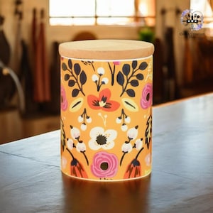 Op de afbeelding: Een gele keramische pot met een bloemenpatroon in tinten roze, rood, wit en zwart. De pot heeft een houten deksel.