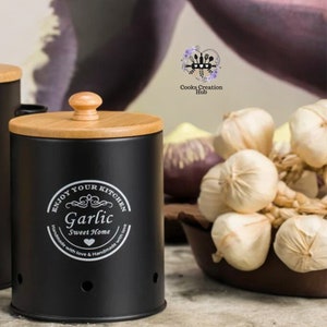 Puede incluir: Un recipiente negro para guardar ajos con tapa y pomo de madera. El recipiente tiene las palabras "Enjoy Your Kitchen" y "Garlic Sweet Home" impresas. Un cuenco con bulbos de ajo en el fondo.