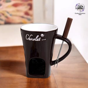 Può includere: Una tazza in ceramica marrone con un manico bianco e la scritta "Chocolat..." sul lato. La tazza ha un foro sul fondo per sciogliere il cioccolato. Viene fornita con una forchetta di metallo per intingere.