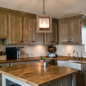 May include: A rustic kitchen with wooden cabinets and a large wooden island. A square, wooden pendant light hangs above the island. The light fixture has a glass enclosure and a warm, inviting glow. The kitchen has a farmhouse style.