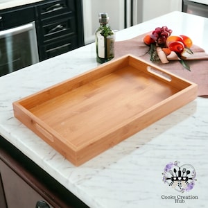 Puede incluir: Una bandeja rectangular de servir de bambú natural con dos asas. La bandeja está vacía y se encuentra sobre una encimera de mármol blanco.