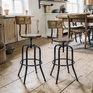 May include: Two industrial-style bar stools with black metal frames and wooden seats and backs. The seats are round and adjustable. The backs are rectangular. The stools are on a wooden floor in a kitchen setting.