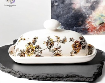 Könnte beinhalten: Eine weiße Keramik-Butterdose mit einem Blumendesign in Gelb-, Braun- und Grüntönen. Die Dose hat einen Deckel und steht auf einem grauen Schiefer-Serviertablett.