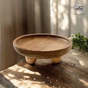 以下が含まれることがあります： 3本の丸い脚が付いた丸い木製サービングトレイ。トレイは天然木で作られており、薄い茶色をしています。