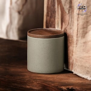 May include: A light gray ceramic jar with a wooden lid. The jar is sitting on a wooden surface.