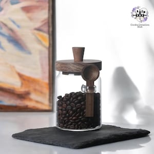 Pode incluir: Um frasco de vidro com tampa de madeira e uma colher de madeira. O frasco está cheio de grãos de café. O frasco está sobre uma superfície de ardósia preta.