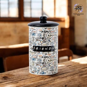 Op de afbeelding: Witte keramische pot met een zwart deksel. De pot is versierd met een zwart-witte illustratie van verschillende scènes en personages uit de tv-serie "Friends". De tekst "FRIENDS" is in zwart op de pot gedrukt.