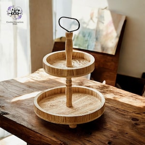 Può includere: Un portavivande in legno a due livelli con una maniglia in metallo nero. Il portavivande ha una finitura in legno naturale ed è perfetto per esporre spuntini, dessert o altri oggetti.