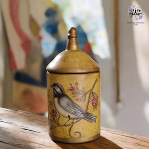 Puede incluir: Un tarro de cerámica amarillo con tapa dorada y un diseño pintado de un pájaro azul posado en una rama con flores rosas. El tarro es un recipiente de almacenamiento decorativo.
