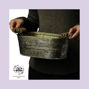 Puede incluir: Un macetero ovalado de metal desgastado con asas de madera. El macetero tiene las palabras "Heartful Garden Since 1972" grabadas en relieve.