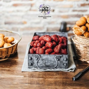 Może przedstawiać: Metalowa taca z uchwytem wypełniona suszonymi czerwonymi daktylami. Taca stoi na drewnianym stole z białym obrusem pod spodem.