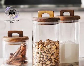 May include: Three glass canisters with wooden lids. The canisters are filled with pistachios, cinnamon sticks, and white powder. The canisters are on a marble countertop.
