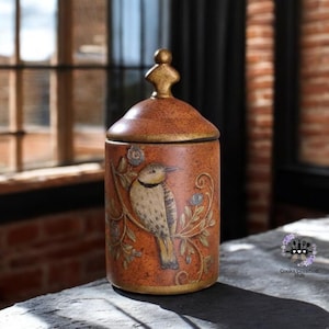 Könnte beinhalten: Dekorative Keramikdose mit Deckel, verziert mit einem handgemalten Vogeldesign auf braunem Hintergrund. Die Dose hat eine goldfarbene Spitze und einen Sockel mit floralen Akzenten. Ein dekorativer Artikel für Küche oder Wohnbereich.