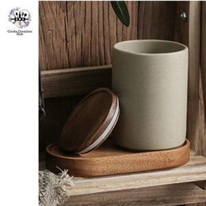 Puede incluir: Un tarro de cerámica beige claro con tapa de madera y una bandeja de madera. El tarro está sentado en la bandeja, que está en un estante de madera.