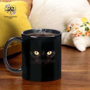 May include: Black ceramic coffee mug with a black cat face design. The cat's eyes are glowing yellow and the mug is filled with coffee.