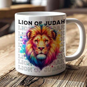 May include: White ceramic mug with a colourful lion illustration and the text "LION OF JUDAH". The lion's mane is a vibrant mix of yellow, orange, blue, and purple. The mug has a white handle and is on a wooden surface.