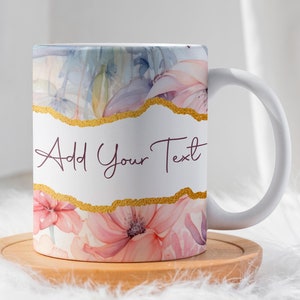 Op de afbeelding: Witte keramische mok met een bloemenontwerp met roze en blauwe aquarelbloemen. De mok heeft een gouden accent en de tekst "Add Your Text" in bordeauxrode schrijfletters.
