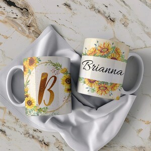 Pu&ograve; includere: Due tazze in ceramica bianca con disegni di girasoli. Una tazza ha una lettera dorata "B" in una ghirlanda di girasoli e verde. L'altra tazza ha il nome "Brianna" in scrittura nera con girasoli intorno.