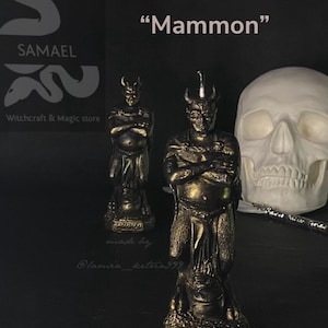 May include: Two bronze-colored statues of a horned figure with crossed arms, labeled "Mammon" candle. A white skull and a metal wand are also present. The image includes the text "SAMAEL Witchcraft & Magic store".