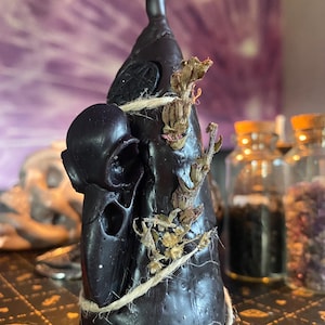 Op de afbeelding: Een zwarte keramische fles met een schedelvormige stop, omwikkeld met touw en versierd met gedroogde bloemen. De fles staat op een zwart-gouden gepatroneerd oppervlak.