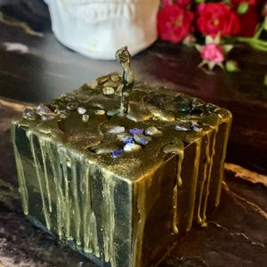 May include: A square black candle with gold wax dripping down the sides. The candle is decorated with small blue and gold stones.
