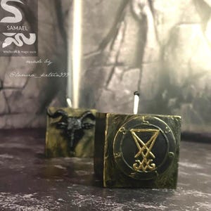 Op de afbeelding: Twee vierkante kaarsen met een donkergroene en gouden afwerking. Eén kaars heeft een geitenkop, de andere een zegel. De kaarsen hebben witte lonten. De afbeelding bevat de tekst "SAMAEL Witchcraft & Magic store".