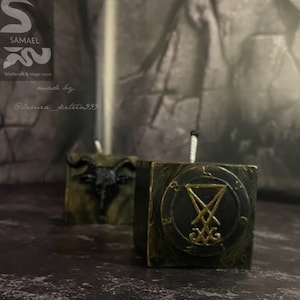 May include: Two black and gold square candles with a pentagram and a goat's head design. The candles are on a dark surface. "Samael Witchcraft & Magic store" and "made by @lamia_ketrin999" are written on the background.