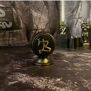 Könnte beinhalten: Ein schwarzes und goldenes Sigil mit dem Symbol "B" in Gold auf schwarzem Hintergrund. Das Sigil steht auf einem kleinen, runden, schwarzen und goldenen Sockel. Das Sigil befindet sich auf einer dunkelbraunen Oberfläche.