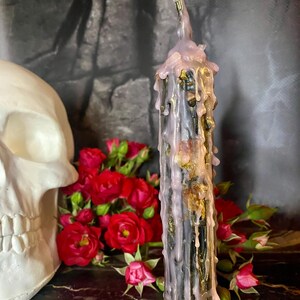 May include: A tall, black candle with dripping lavender wax and gold accents. The candle is next to a white skull and a bouquet of red roses, creating a gothic aesthetic.