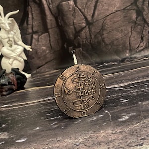 May include: A round, bronze-coloured metal sigil with a complex design featuring a snake, a cross, and other symbols. The sigil is surrounded by a circle with the letters "A.S.M.U.D.E.D." inscribed around it.