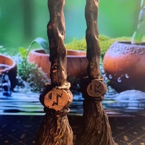 Puede incluir: Dos velas de escoba marrones y doradas con un rayo y un símbolo de runa en los mangos. Las velas están sobre una superficie con estampado negro y dorado.