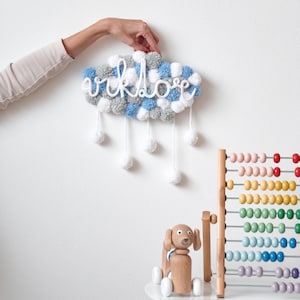 May include: A white cloud-shaped pom-pom wall hanging with the word "vklove" spelled out in white yarn. The cloud has blue and grey pom-poms and white yarn strings hanging down. A wooden toy dog and a rainbow-coloured abacus are on a white table in the background.