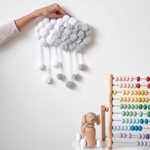 May include: A white and grey pom-pom cloud mobile with white pom-pom raindrops hanging from it. A wooden toy dog and a rainbow abacus are on a white table in the background.