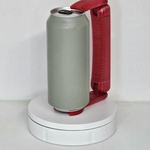 May include: A gray aluminum can with a red plastic handle and opener. The can is on a white rotating platform. The handle is designed to make it easier to hold and open the can.