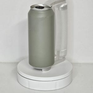 May include: A sage green aluminum can with a silver top, held by a clear plastic handle. The can is on a white, circular platform, likely for display or photography. The handle is attached to the can.