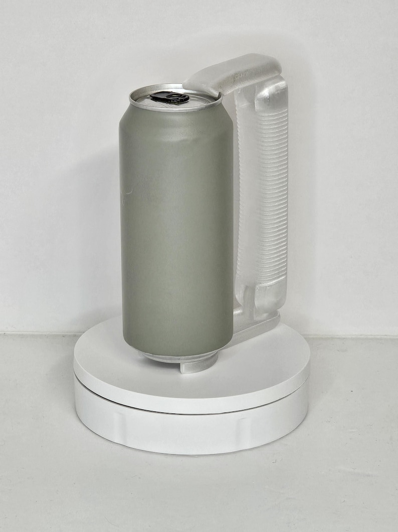 May include: A sage green aluminum can with a silver top, held by a clear plastic handle. The can is on a white, circular platform, likely for display or photography. The handle is attached to the can.