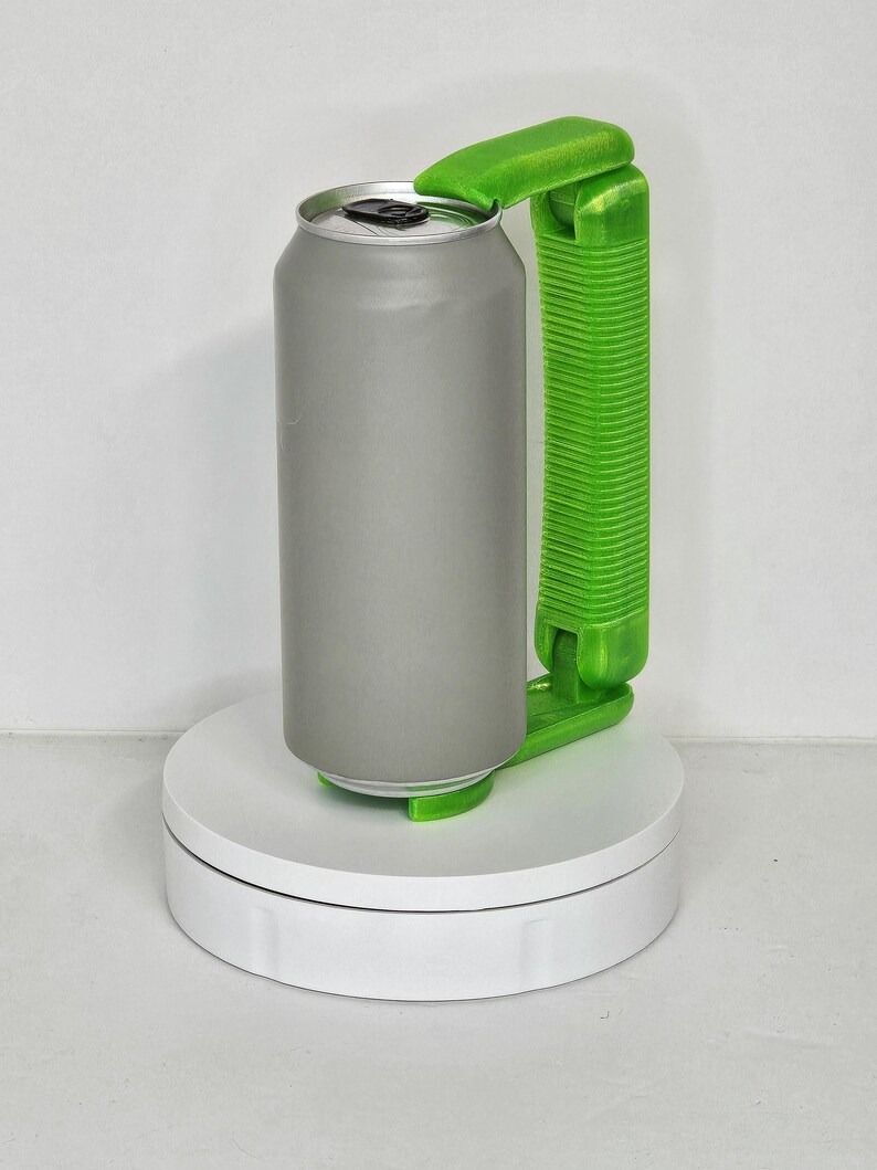 May include: A gray aluminum can with a silver top is held by a bright green can opener. The can opener has a handle and a lever to lift the can's lid. The can and opener are on a white rotating platform.