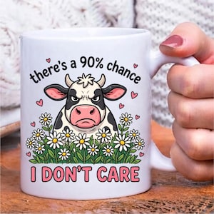 Pode incluir: Uma caneca de cer&acirc;mica branca com o gr&aacute;fico de uma vaca rabugenta em preto e branco rodeada por margaridas. O texto acima da vaca diz there's a 90% chance e o texto abaixo diz I DON'T CARE, acentuado por pequenos cora&ccedil;&otilde;es cor-de-rosa.