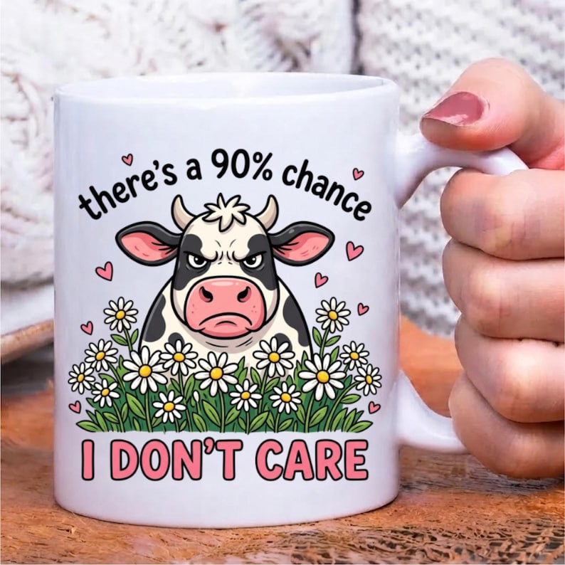 Pode incluir: Uma caneca de cer&acirc;mica branca com o gr&aacute;fico de uma vaca rabugenta em preto e branco rodeada por margaridas. O texto acima da vaca diz there's a 90% chance e o texto abaixo diz I DON'T CARE, acentuado por pequenos cora&ccedil;&otilde;es cor-de-rosa.