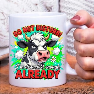Pode incluir: Uma caneca de cer&acirc;mica branca com o gr&aacute;fico de uma vaca rabugenta em preto e branco com chifres. O texto DO NOT DISTURB! I'm disturbed enough ALREADY est&aacute; em letras vermelhas e pretas, rodeado por salpicos de tinta verde e nuvens.