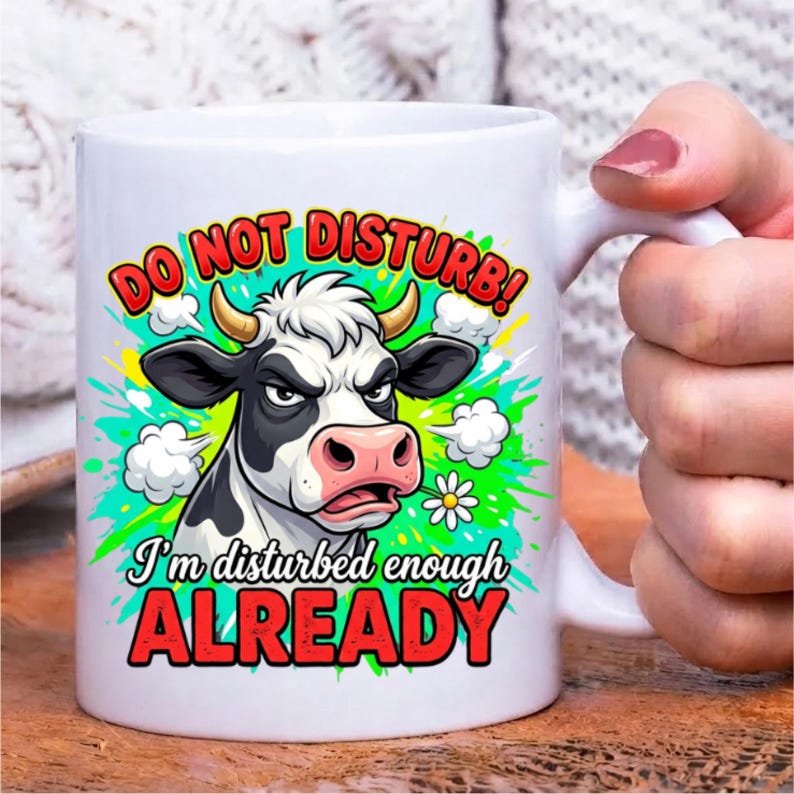 Pode incluir: Uma caneca de cer&acirc;mica branca com o gr&aacute;fico de uma vaca rabugenta em preto e branco com chifres. O texto DO NOT DISTURB! I'm disturbed enough ALREADY est&aacute; em letras vermelhas e pretas, rodeado por salpicos de tinta verde e nuvens.